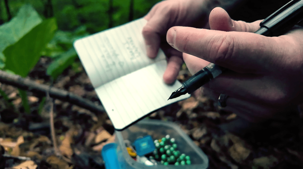 Hands write in a tiny notebook in the woods.