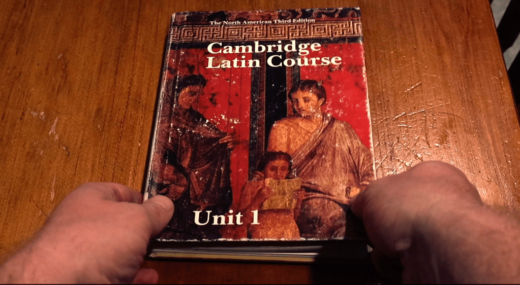 Hands clutch a copy of the Cambridge Latin Course.